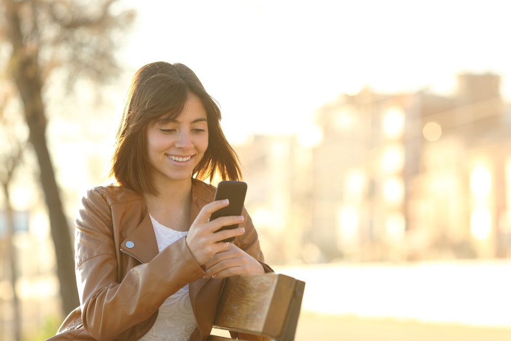 Young woman smiling in the park whilst look at how to unlock her phone to any network.