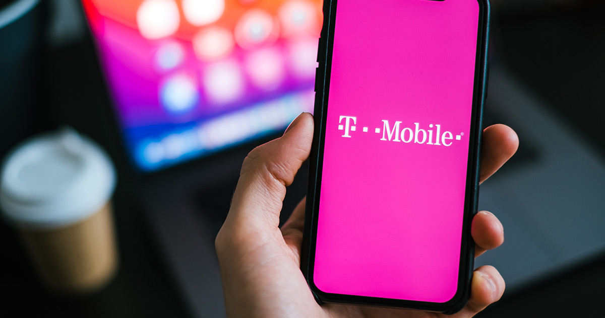 Sosteniendo un teléfono móvil que muestra el logo oficial de T-Mobile en la pantalla de inicio con una computadora portátil en el fondo de la imagen.
