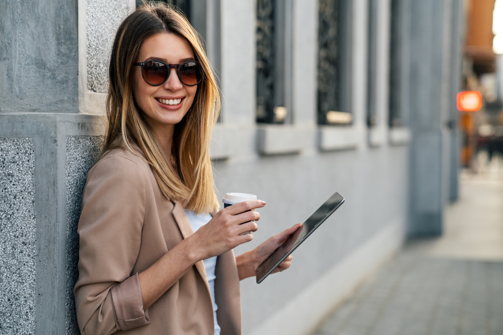 Beautiful blond woman lent against a wall, holding a coffe and her Samsung IMEI checked phone. 